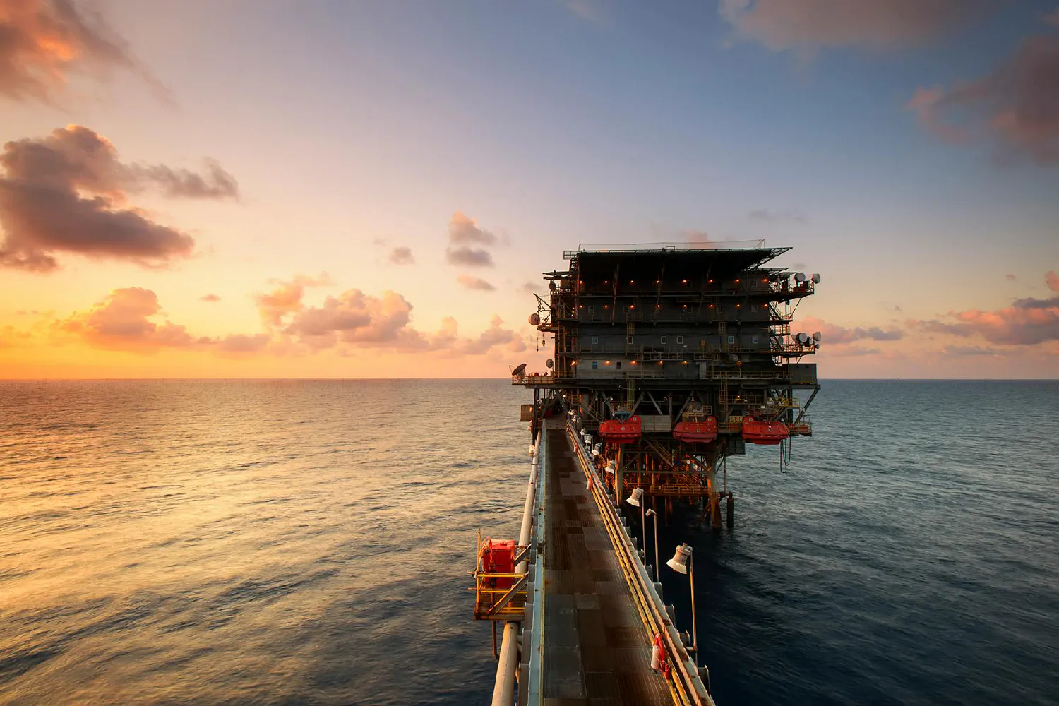 Offshore oil platform at sunset, connected by a metal walkway extending over calm ocean waters, with a colorful sky of orange, pink, and blue hues.